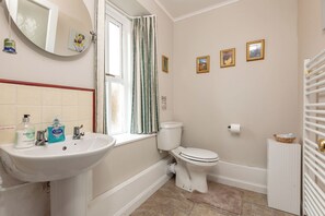 Ein Badezimmer mit weißem Waschbecken, Toilette und einem Fenster mit Vorhängen. Drei Bilder sind in Rahmen an der Wand zu sehen.