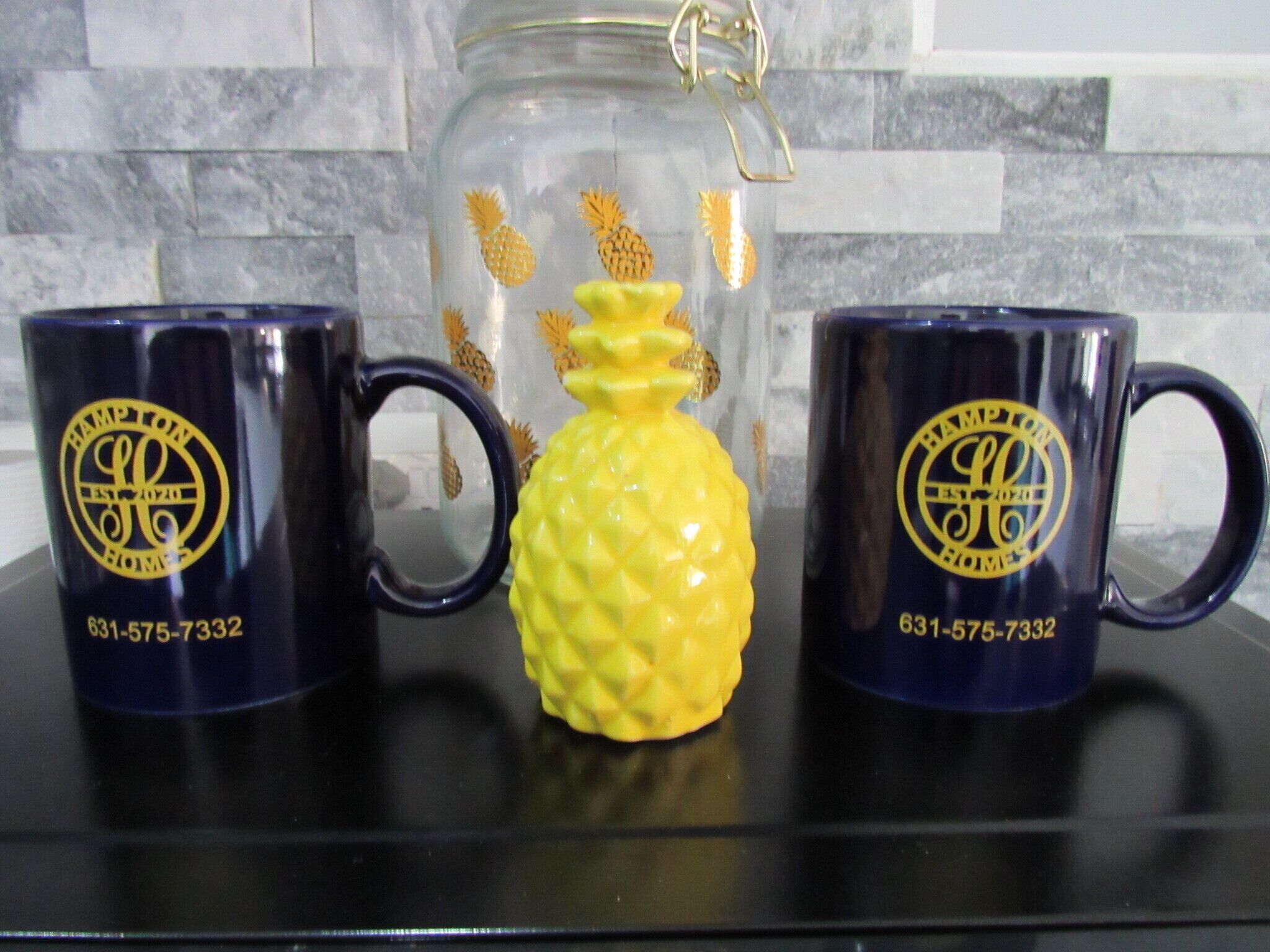 Two dark blue mugs with a logo and phone number, and a yellow pineapple-shaped container on a black countertop.