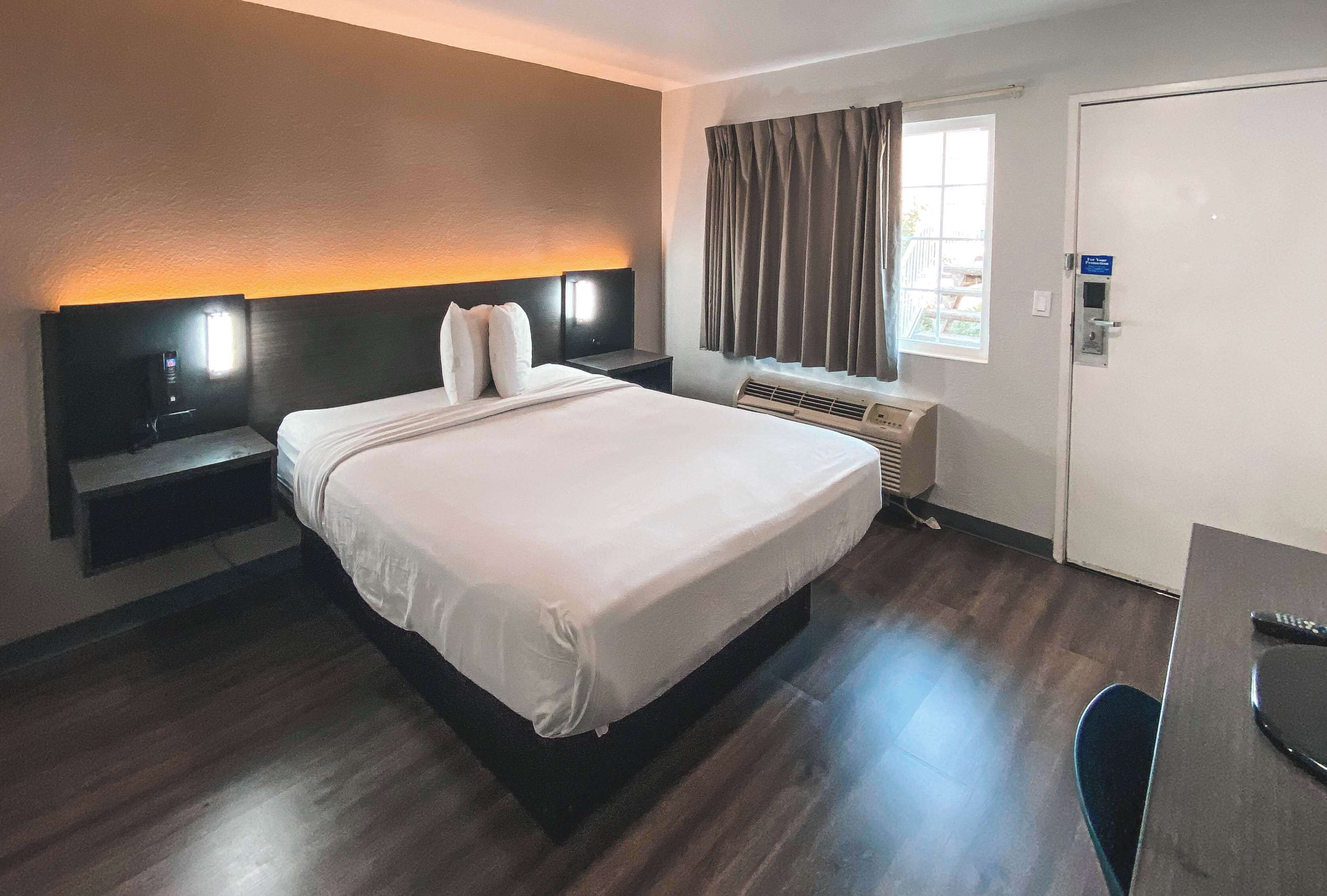 Ein Hotelzimmer mit einem großen Bett, Nachttischen, einem Schreibtisch, einem Sessel, einem Fenster mit Vorhängen, einer Klimaanlage und einer Tür.