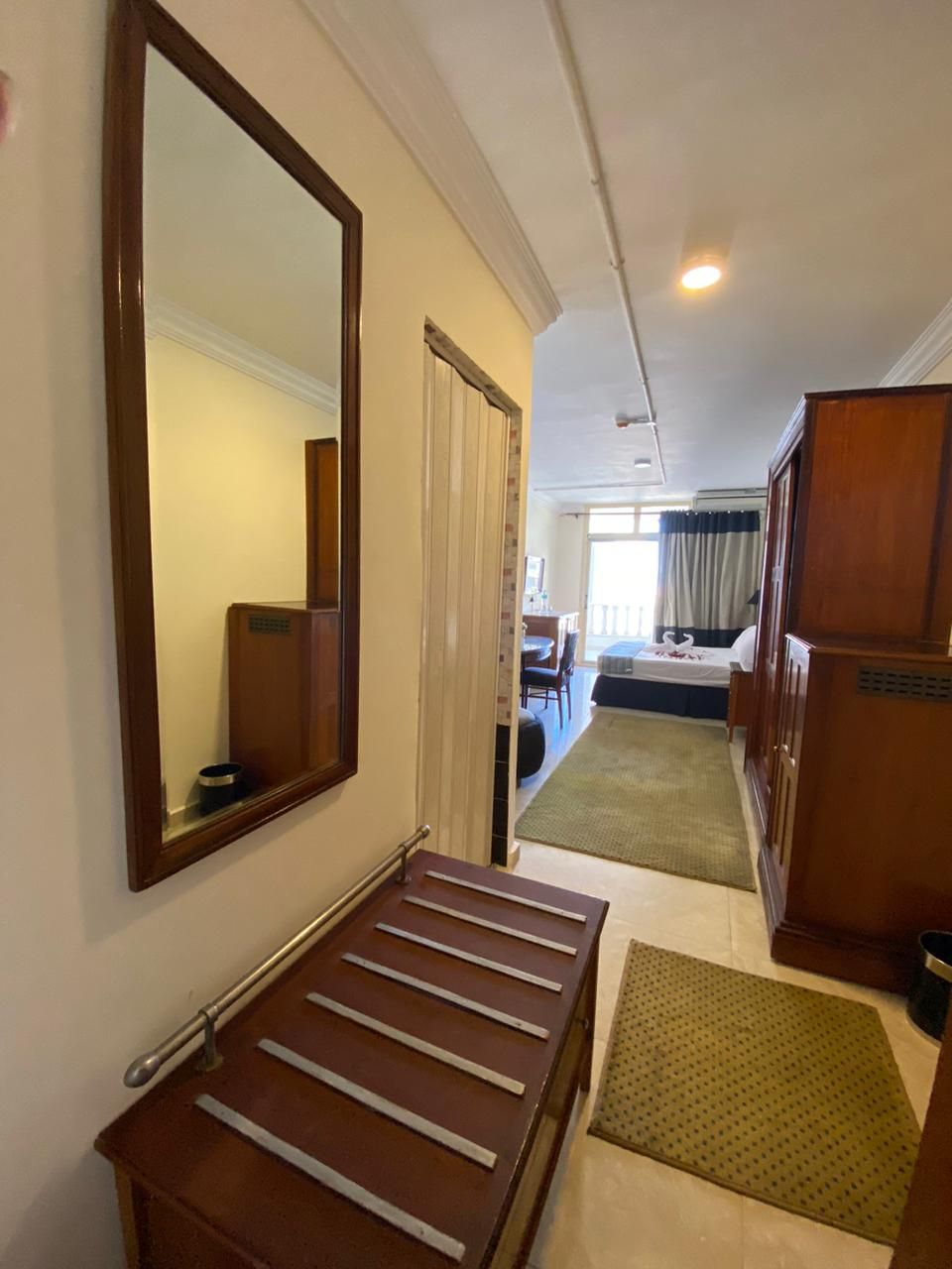 A hotel room with a wooden bench, a large mirror, a bed, a desk, and a small rug.