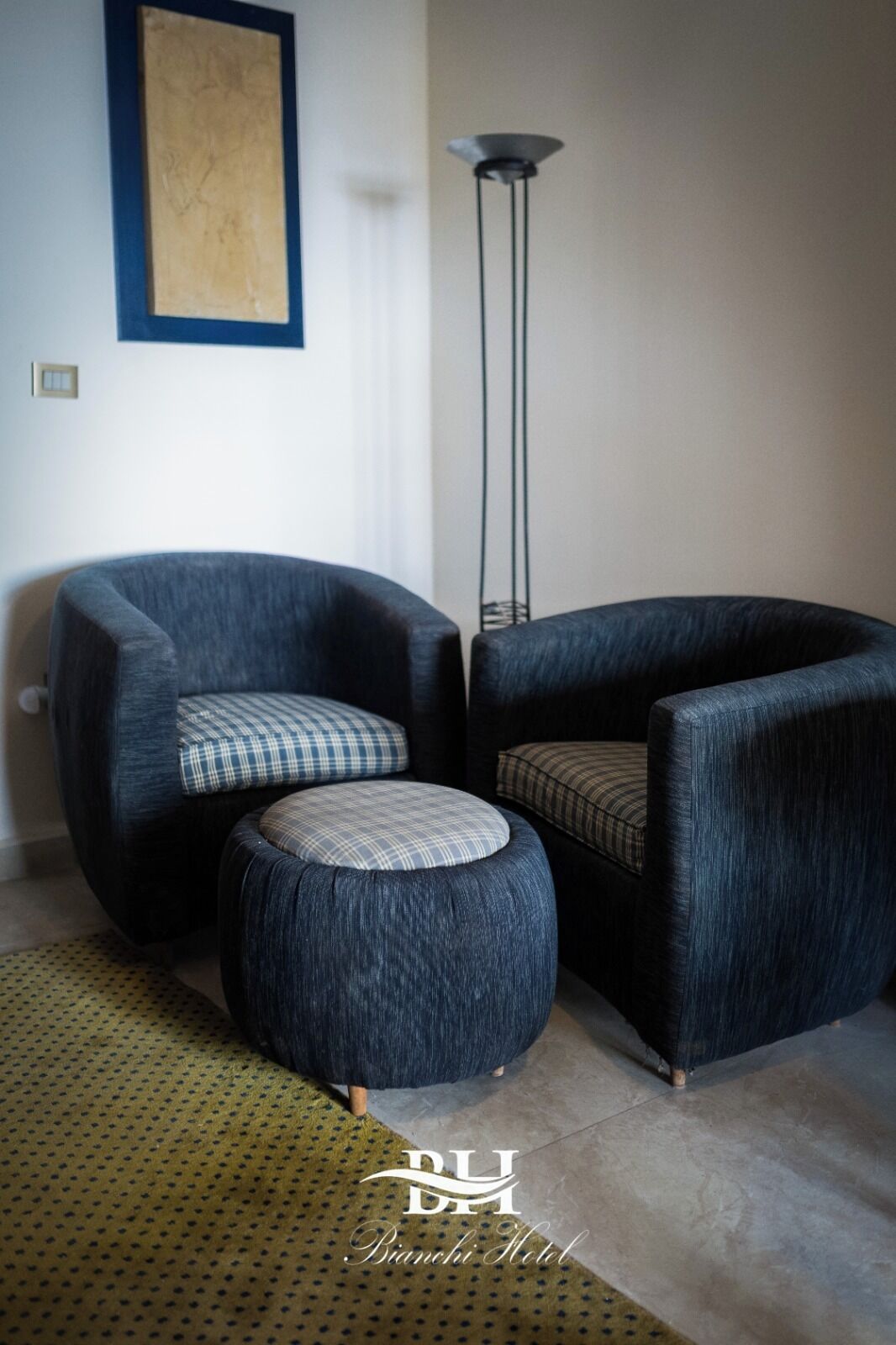 Un salon moderne avec deux fauteuils bleu foncé, l’un orné d’un coussin à carreaux et l’autre d’un coussin uni. Un petit pouf à carreaux est placé entre les deux fauteuils. Un grand lampadaire se dresse dans un coin, et un tableau est accroché au mur. La pièce présente une palette de couleurs neutres, avec un tapis à motifs verts sur le sol.