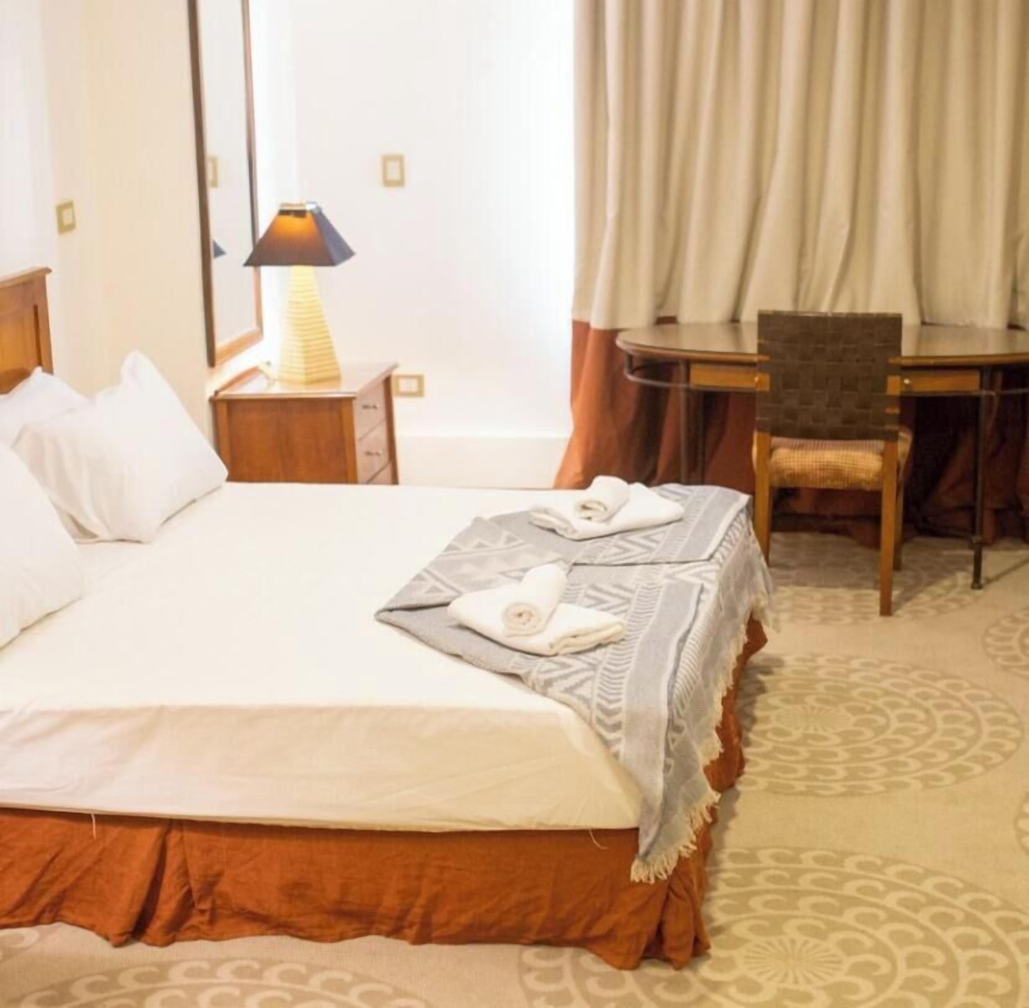 Quarto de hotel com cama, escrivaninha, cadeira, luminária e janela com cortinas.