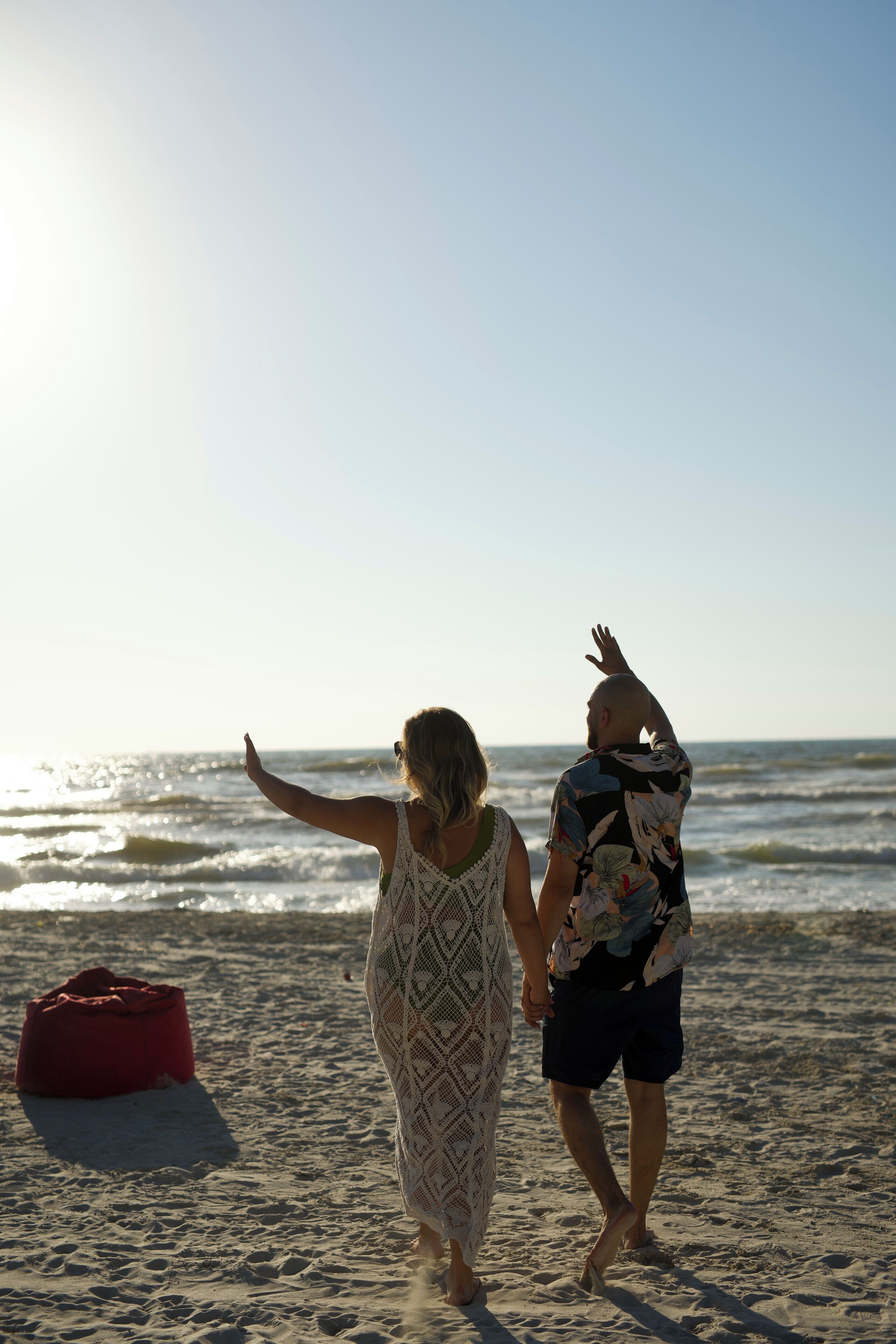 Dos personas caminando sobre una playa arenosa con una bolsa roja y un cielo despejado.