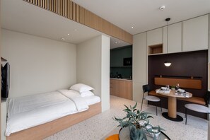 Una habitación de hotel moderna con una cama, una mesa de comedor con sillas, una pequeña cocina y una zona de estar con un sofá y una planta.