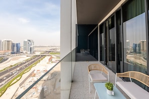 A balcony with a view of a cityscape, featuring modern buildings and a road. There are two wicker chairs and a small table with a plant.