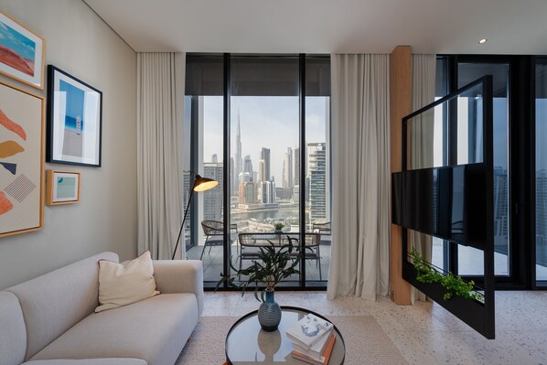 Una sala de estar moderna con un sofá, una mesa de centro con un florero y libros, un televisor sobre un mueble bajo y un ventanal con vistas a la ciudad.