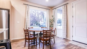 Un comedor con una mesa y sillas de madera, un refrigerador de acero inoxidable y vista a una calle arbolada a través de amplios ventanales.