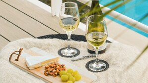 Un balcon avec vue sur la piscine, une bouteille de vin blanc, deux verres, une planche de fromages accompagnée de raisins et une petite figurine.