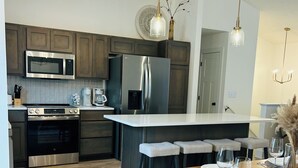 A modern kitchen with stainless steel appliances, a central island with bar stools, and pendant lighting. - New construction, Sunset views  (Lincoln)