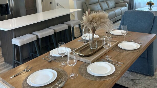 A modern dining area with a wooden table set for four, a kitchen in the background, and a living area with a sofa and a blue chair. - New construction, Sunset views  (Lincoln)