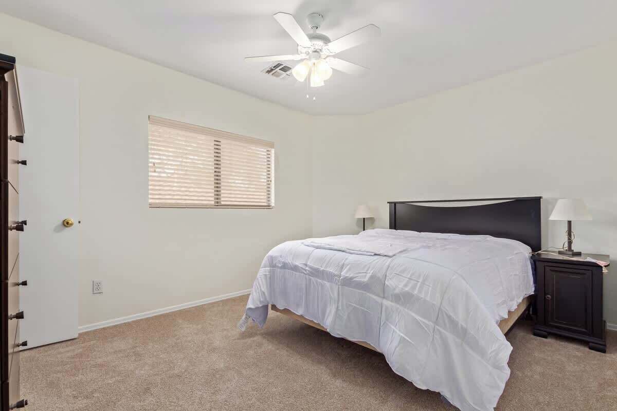 A bedroom with a bed, a ceiling fan, a window with blinds, and a nightstand with a lamp.