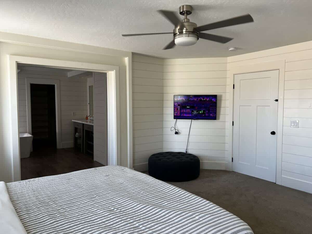 A bedroom with a bed, a ceiling fan, a TV mounted on the wall, and a door.