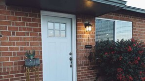 A brick house with a white front door, a small potted plant, and a red flowering bush.