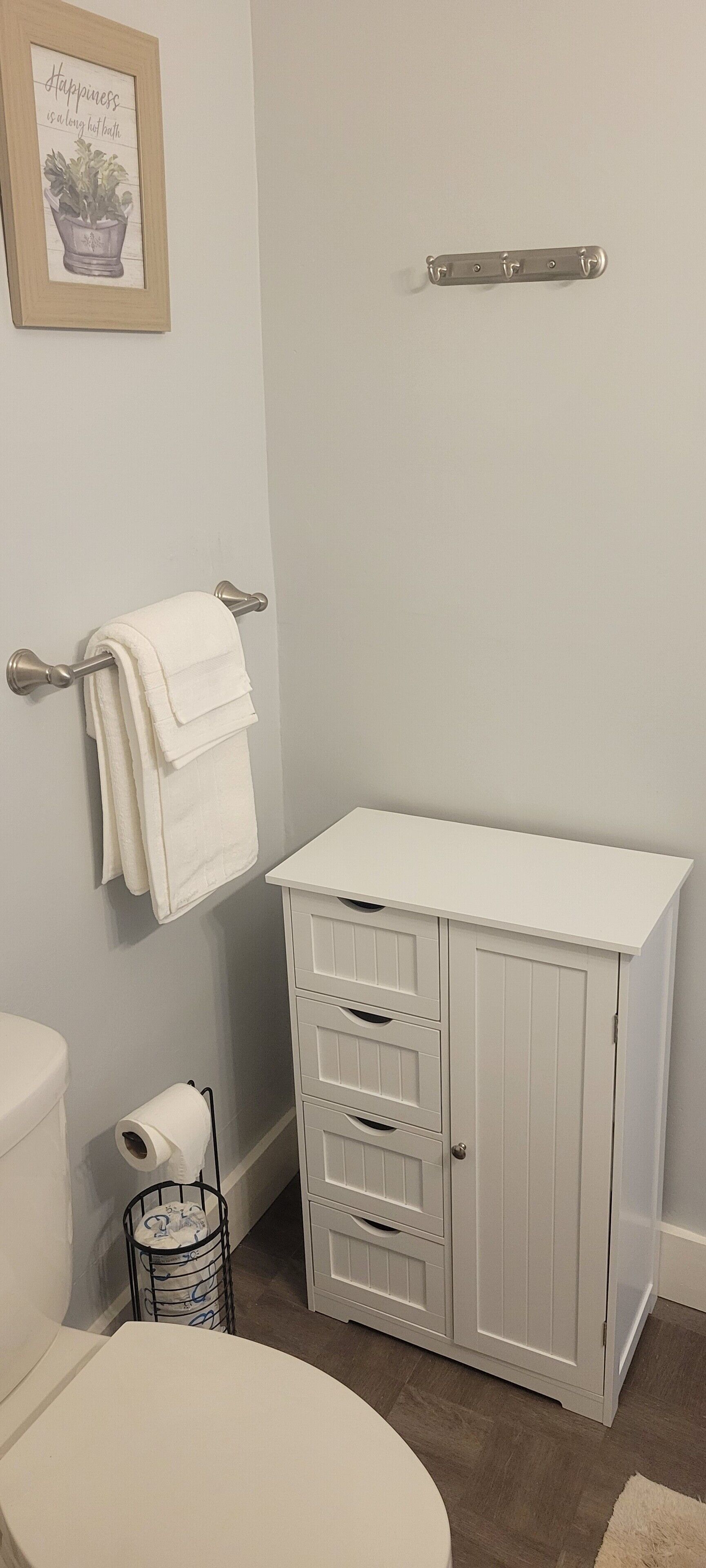 Un baño con inodoro blanco, un perchero con toalla blanca, un mueble con cajones y una cita enmarcada en la pared.