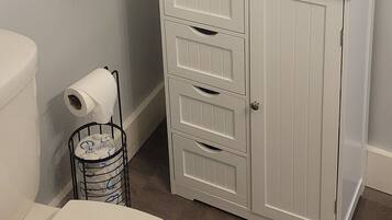 Un baño con inodoro blanco, un perchero con toalla blanca, un mueble con cajones y una cita enmarcada en la pared.