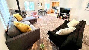 A living room with a sofa, armchair, and coffee table. There is a kitchen area with cabinets and a TV in the background.