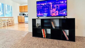 A living room with a flat-screen TV on a black stand, a kitchen in the background, and a dining area with wooden furniture.