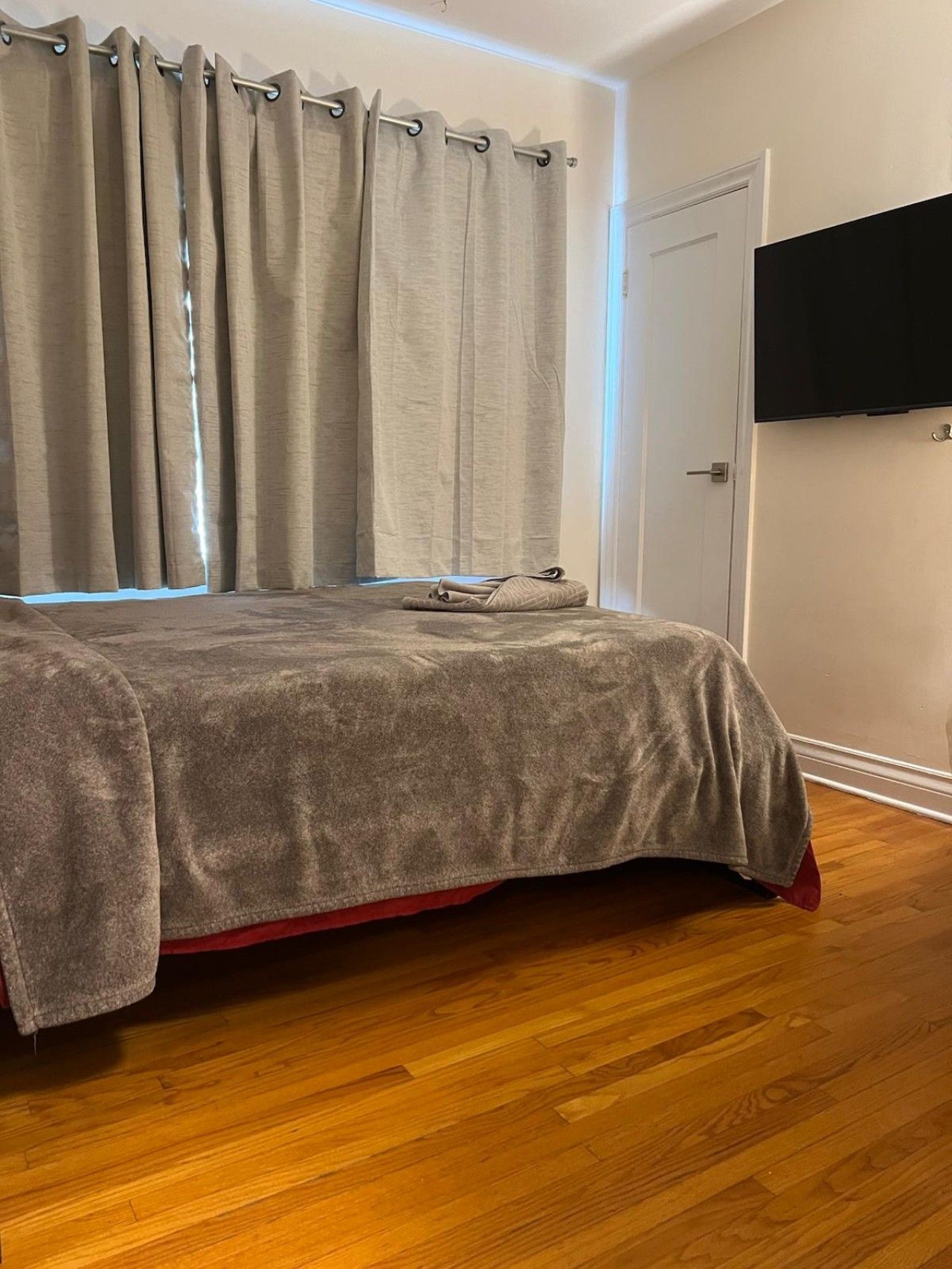 A bedroom with a bed, curtains, a flat-screen TV, and a wooden floor.