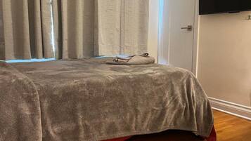 A bedroom with a bed, curtains, a flat-screen TV, and a wooden floor.