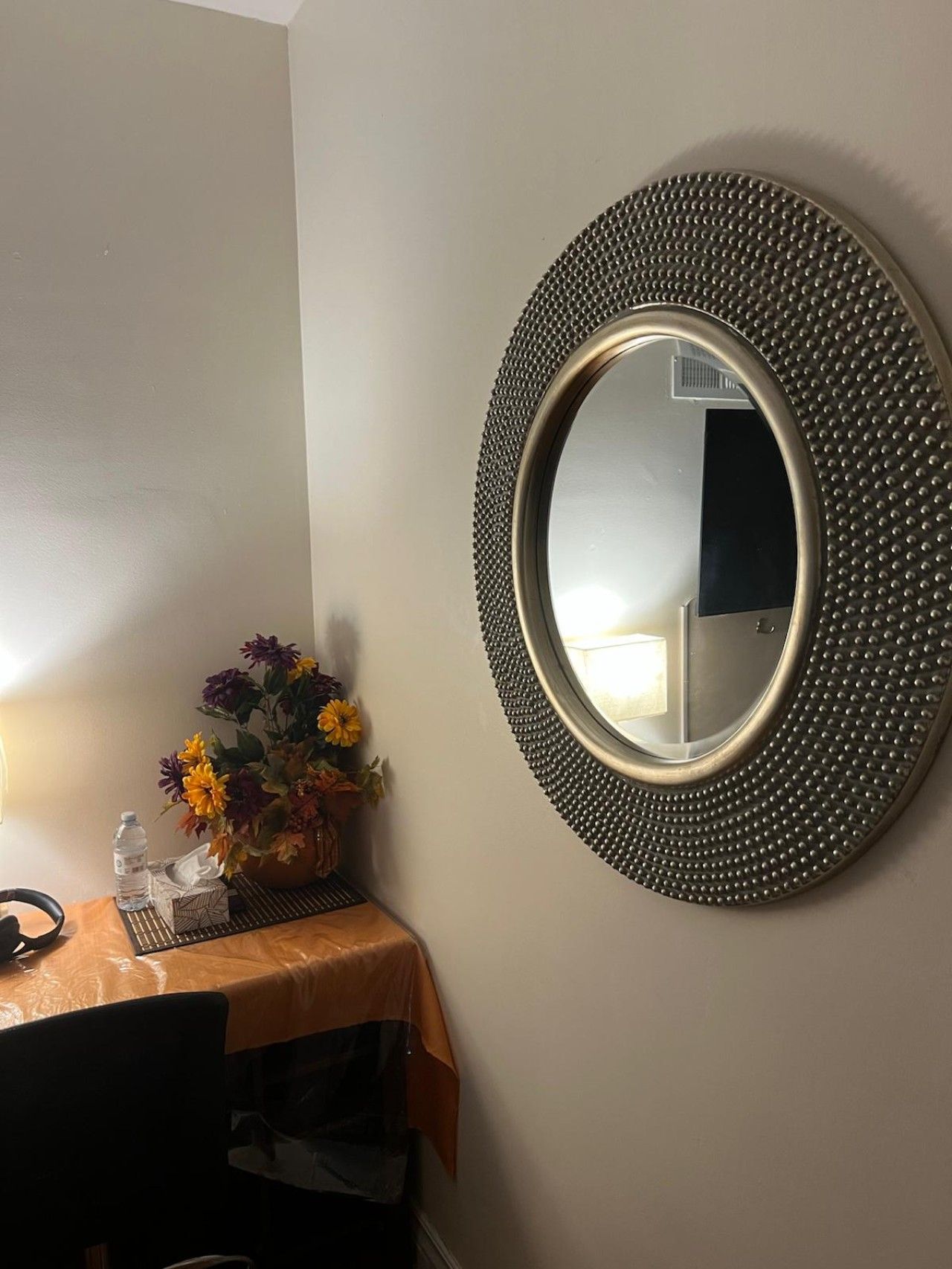 A round mirror with a textured frame on a wall, a table with a plastic tablecloth, a vase with flowers, and a bottle of water.