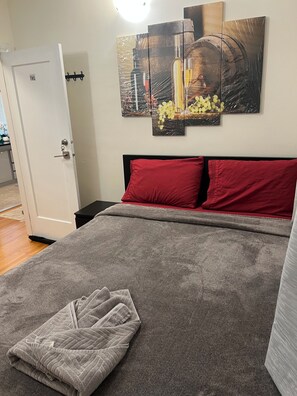 Un dormitorio con una cama, un almohadón rojo, una manta gris y una obra de arte en la pared.