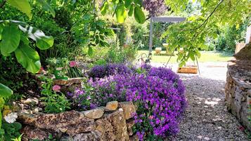 Ein Garten mit Steinmauer, Kiesweg und verschiedenen Pflanzen, darunter auch violette Blumen.