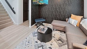 Una sala de estar moderna con una escalera, un televisor de pantalla plana en la pared, un sofá gris con cojines estampados y una mesa de centro redonda con revistas.