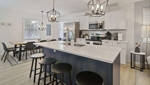 Una cocina moderna con armarios blancos, una isla central con taburetes, electrodomésticos de acero inoxidable e iluminación colgante.