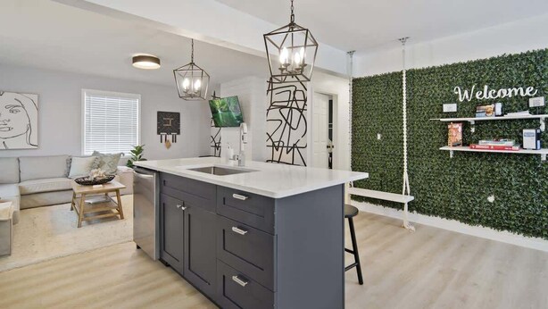Una cocina moderna con una isla central, luces colgantes y una pared verde con un letrero que dice 'Welcome'.