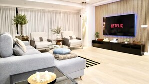 En moderne stue med et stort fladskærms-tv, der viser 'NETFLIX', en grå sofa, to lænestole, et sofabord med stearinlys og en dekorativ plante.