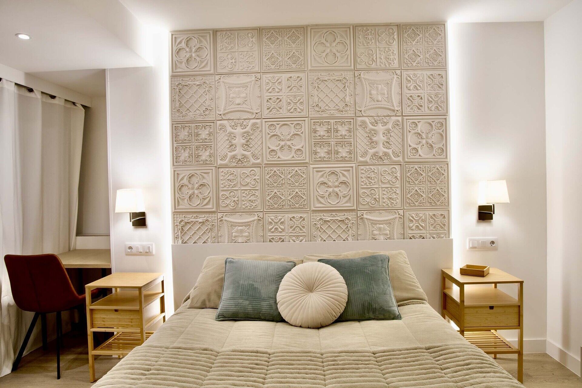 A bedroom with a decorative headboard, a bed with multiple pillows, a wooden nightstand, a desk, and a chair.