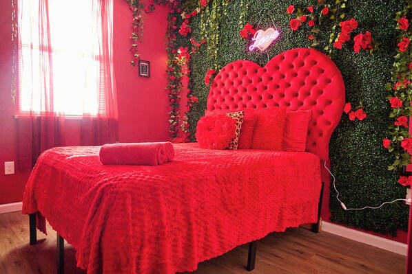 Une chambre à coucher aux couleurs rouge, avec une tête de lit capitonnée, un néon en forme de cœur et un mur verdoyant orné de fleurs rouges.