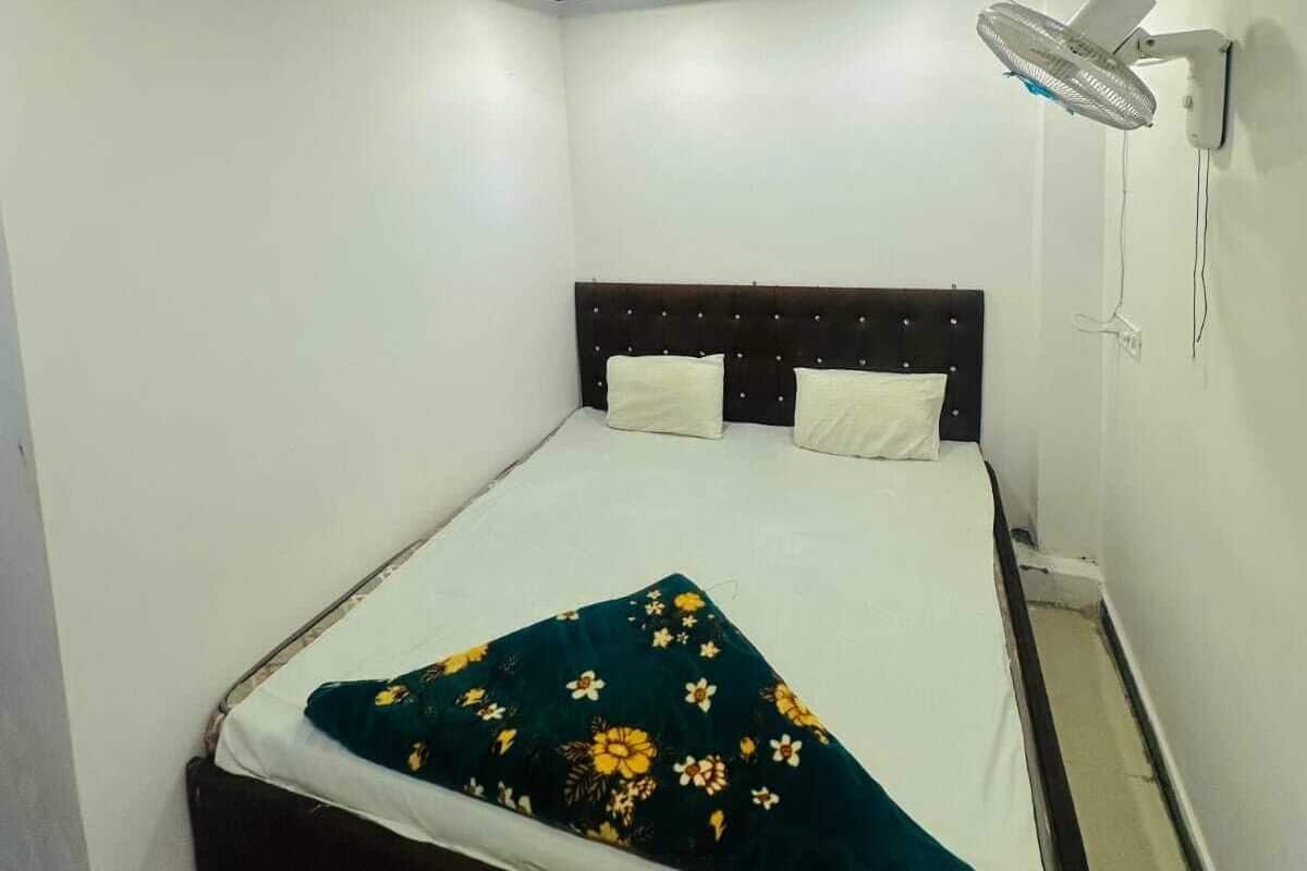Une petite pièce propre, avec un lit simple, un ventilateur fixé au mur et un luminaire au plafond.