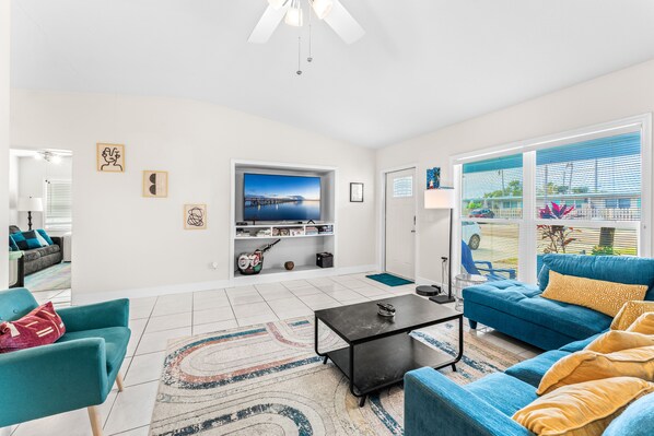 A modern living room with a large TV, teal and blue furniture, and a view of the outdoors through large windows. - The Pelican Cove- <4 min walk to Cocoa Beach (Cocoa Beach)