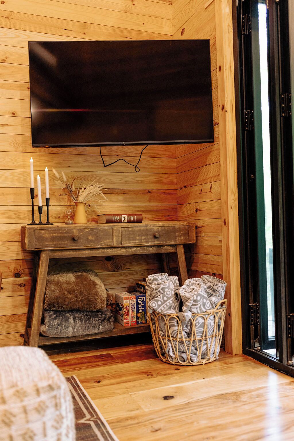 Un intérieur en bois agrémenté d’un téléviseur à écran plat fixé au mur, d’un banc en bois avec des bougies, d’un vase contenant des plantes séchées et d’un panier rempli de serviettes pliées.