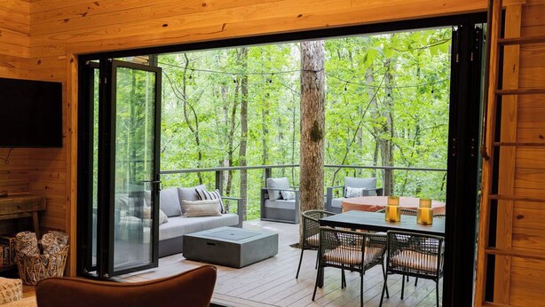 A wooden cabin with a glass door leading to an outdoor deck. Inside, there is a sofa, a table with candles, and wicker chairs. - Beech Retreet Treehouse Atlanta  (Fairburn)