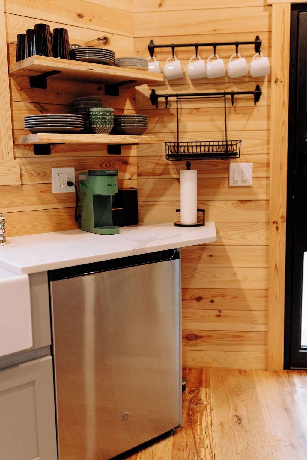 A compact kitchen with wooden walls, a stainless steel dishwasher, a coffee maker, and a hanging mug rack.