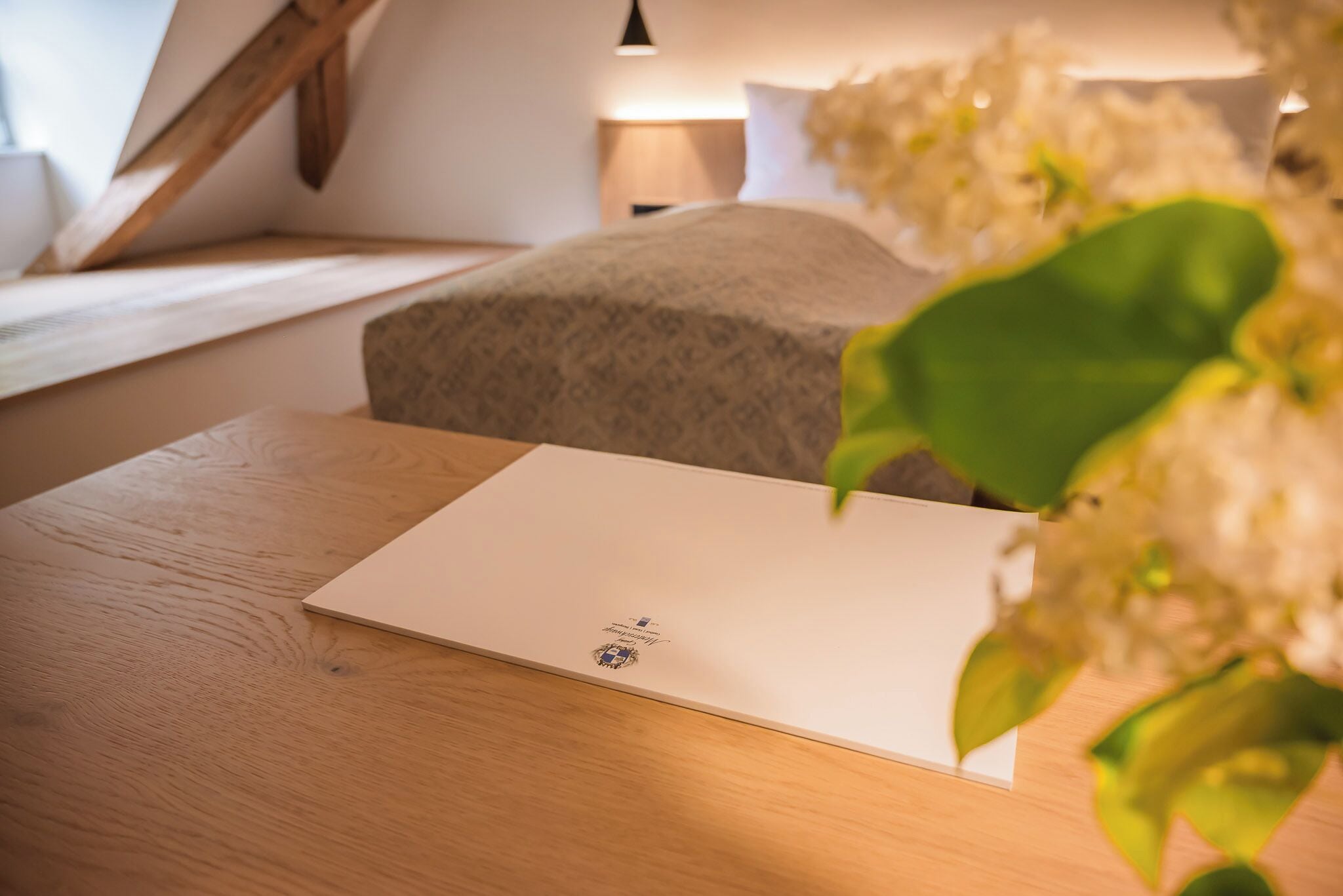 Una habitación ordenada con una mesa de madera, una cama con cubrecolcha gris estampada y una lámpara al fondo.