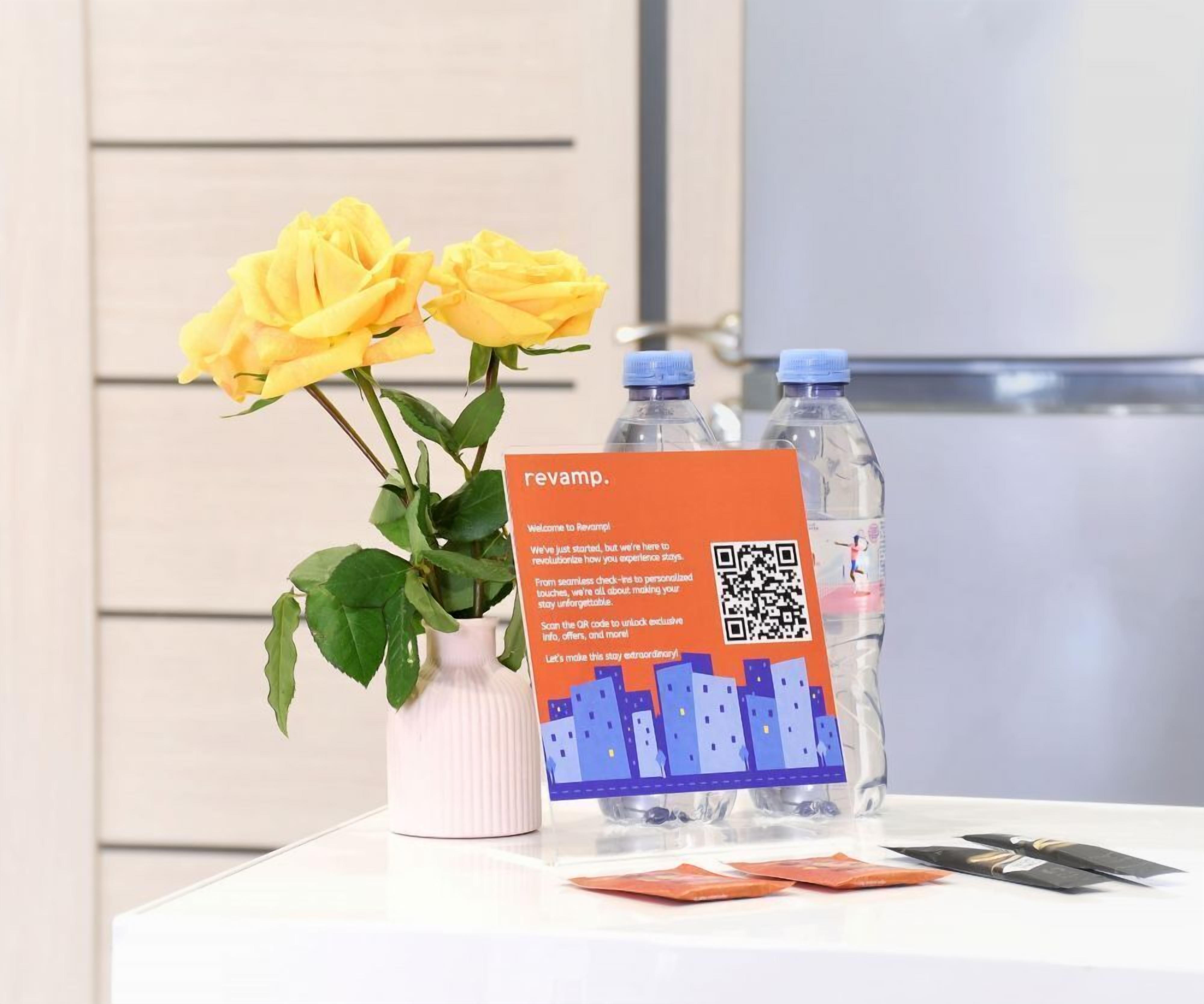 Une carte de bienvenue pour l’hôtel Revamp, un code QR et deux bouteilles d’eau sont posés sur une table, à côté d’un vase rempli de roses jaunes.