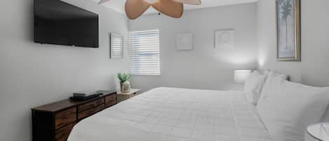 A bedroom with a bed, a TV mounted on the wall, a ceiling fan, and a window with blinds.