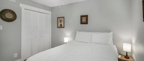 A bedroom with a bed, two bedside lamps, a ceiling fan, a white closet, and two framed pictures on the wall.