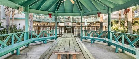 A covered picnic area with a wooden table and benches, surrounded by a green fence and palm trees.