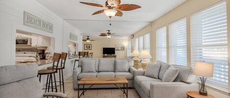 A spacious living room with a sofa, coffee table, and a ceiling fan. There is a kitchen area with a microwave and a sand bar sign.