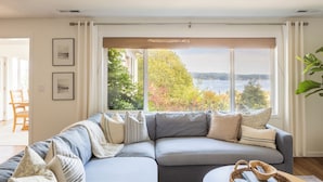 Una sala de estar moderna con un ventanal grande, un sofá seccional gris y vistas a un lago y árboles.