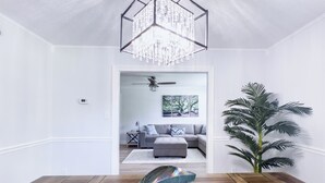 Una sala de estar moderna con piso de madera, una lámpara de cristal, un sofá gris y una planta grande en maceta.