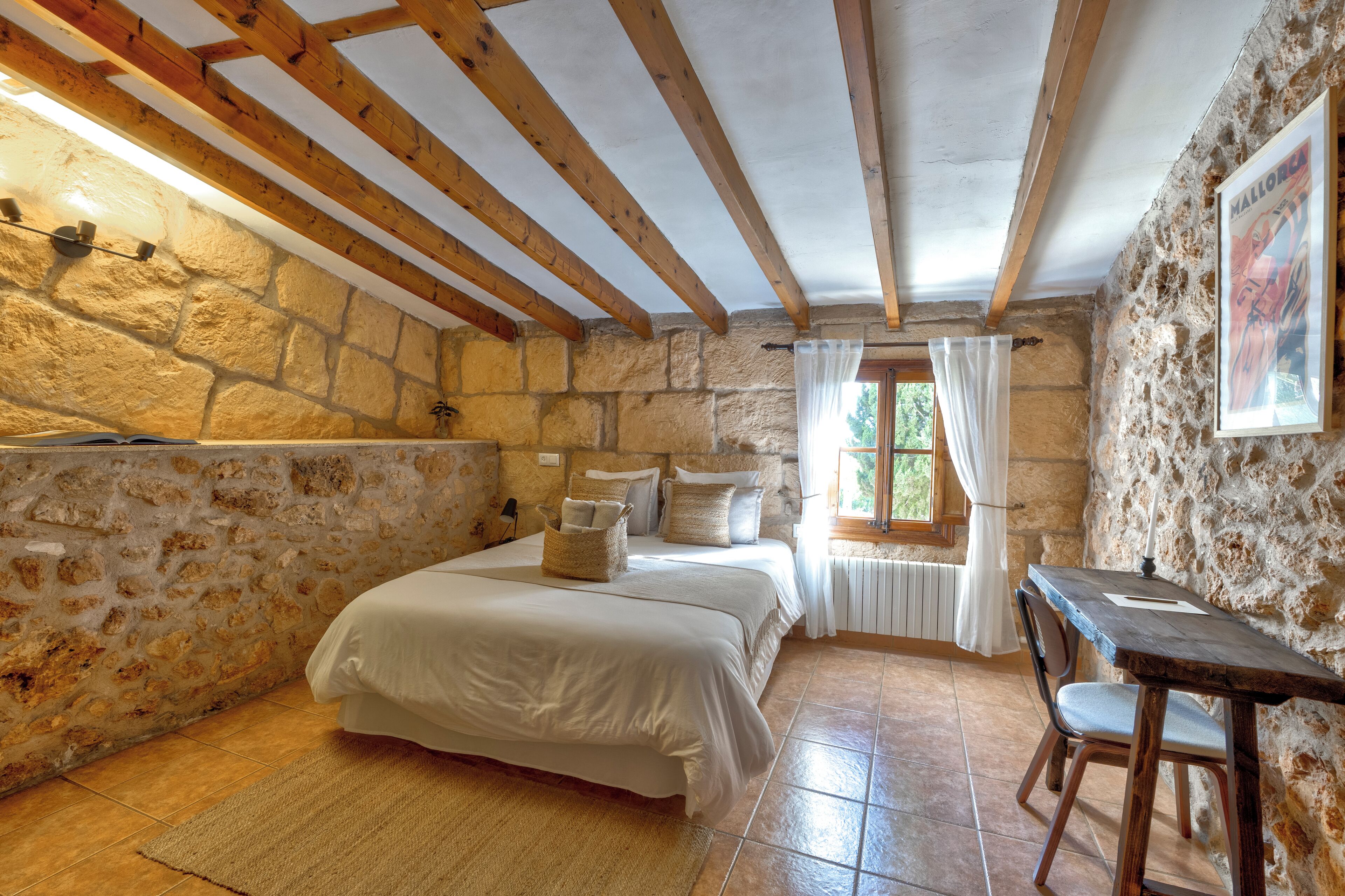 Una habitación rústica con pared de piedra, vigas de madera, una cama con almohadas, una mesa y una silla.