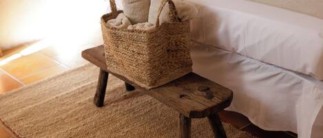 A small wooden stool with a woven basket on a textured rug in a room with a bed and white bedding.