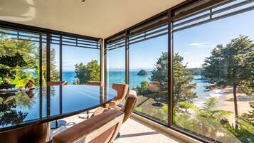 A modern dining area with a glass table and chairs, overlooking a scenic coastal landscape with trees and a view of the sea.