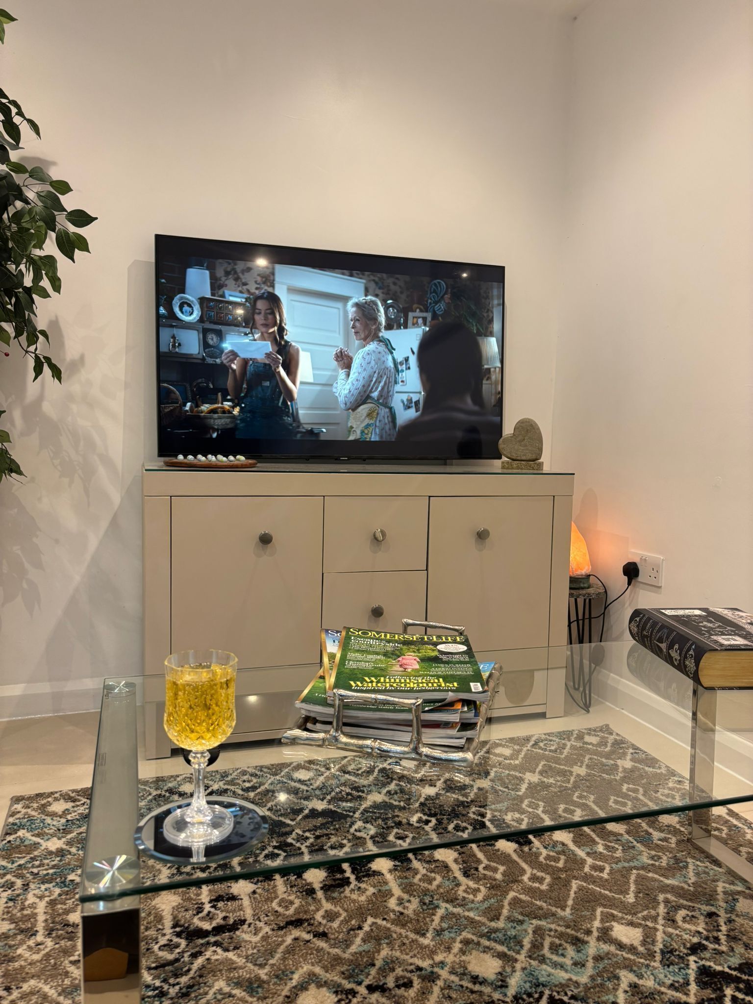Ein Wohnzimmer mit einem Flachbildfernseher an einer Kommode, einem Glastisch mit Zeitschriften und einer Kerze sowie einem gemusterten Teppich.