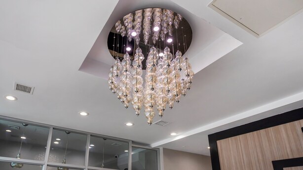 A modern chandelier with multiple tiers of crystal and lights, a recessed ceiling light, and a glass partition with a view. - Red Roof PLUS+ Brooklyn - Flatbush Ave (Brooklyn)